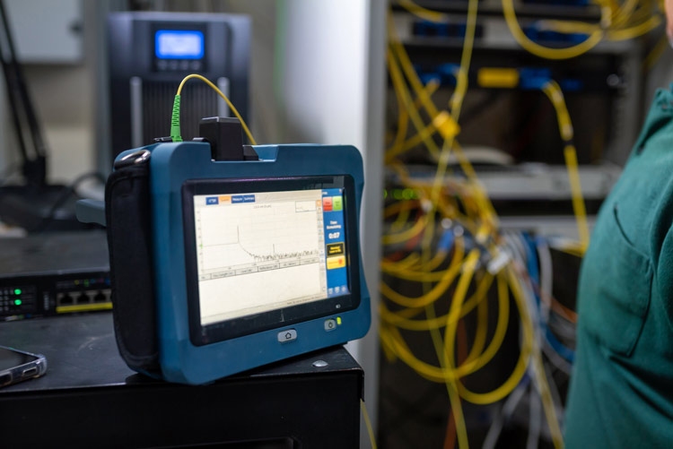 The image shows a professional network tester, a blue portable measuring device with a digital screen used to test the signal strength and operational readiness of fiber optic cables.